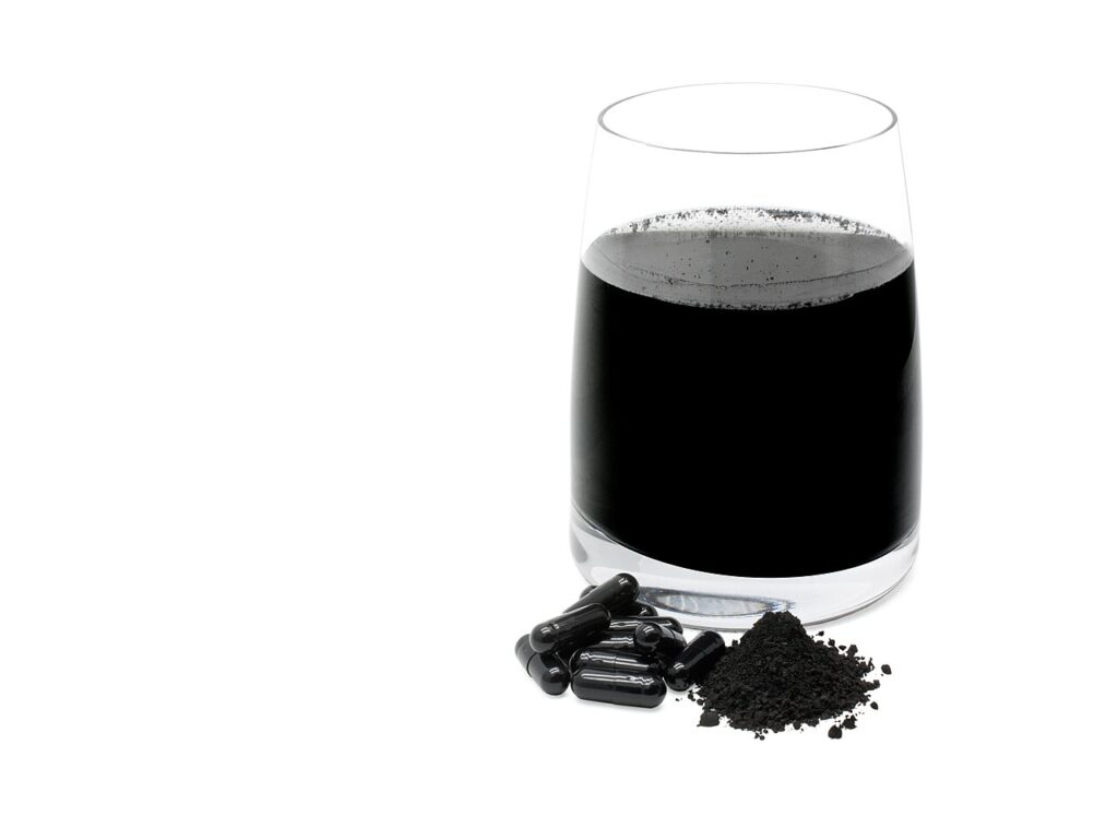 A glass of black activated charcoal liquid next to charcoal tablets and loose powder