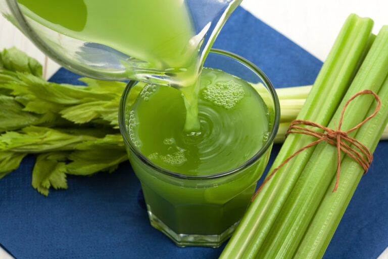 Green celery juice being poured into a glass beside fresh celery stalks
